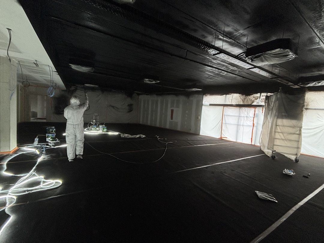 Peintre en combinaison projetant de la peinture noire au pistolet airless sur le plafond technique d’un grand local professionnel, sols et fenêtres protégés